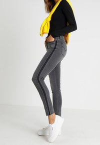 Jeans ajustados grises con rayas laterales negras, con una cintura alta y largo hasta el tobillo. Combinados con una blusa negra de manga larga y zapatillas blancas.