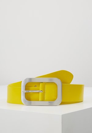 Ceinture en cuir jaune avec une grande boucle rectangulaire argentée reposant sur une surface blanche sur fond uni.
