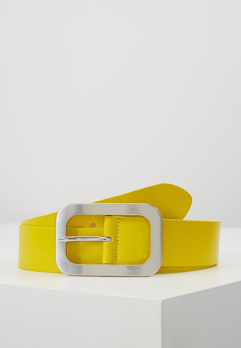Ceinture en cuir jaune avec une grande boucle rectangulaire argentée reposant sur une surface blanche sur fond uni.