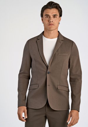 Blazer marrón hecho de tejido texturizado, con un cierre de un solo botón, solapas en pico y dos bolsillos frontales. Combinado con una camisa blanca.
