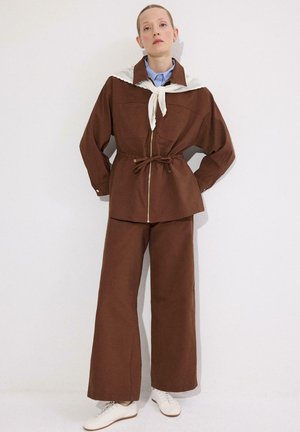 Žena oblečená v hnedých širokých nohaviciach a zodpovedajúcej bunde na zips s pleteným bielym svetrom zviazaným okolo ramien, stojaca pri bielej stene.