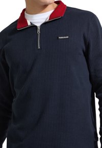 Maglione blu navy a coste con zip al collo, caratterizzato da un colletto rosso a contrasto e un accento bianco, etichetta con logo sul petto.