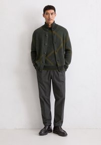 Dark green knitted cardigan with yellow diamond pattern, buttoned, paired with gray tailored trousers and black lace-up shoes.
