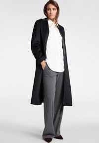 Lungo cappotto nero sopra una camicia bianca con colletto, pantaloni grigi a gamba larga e scarpe marrone scuro. Il cappotto presenta una texture liscia e un design su misura.