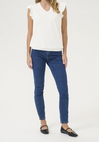 Kobieta ubrana w białą koszulę bez rękawów z falbanami, niebieskie obcisłe dżinsy i czarne buty Mary Jane, stojąca na jednolitym tle.