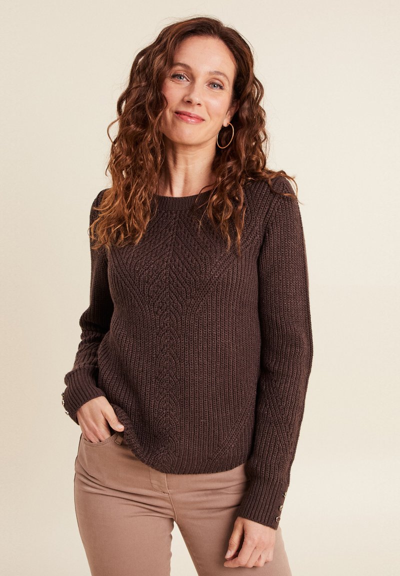 Brauner gestrickter Pullover mit einem strukturierten Rautenmuster, langen Ärmeln und Knopfdetails an den Bündchen, getragen über beigen Hosen.