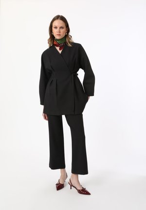 Black wrap jacket with wide sleeves, belted waist, paired with straight black trousers and deep red pointed-toe slingbacks.