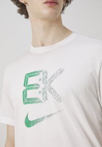 Hvit bomull t-skjorte med et grønt og sprukket grafisk design av "EK" og en stilisert Nike swoosh-logo i lignende tekstur.