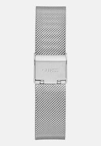 Banda de reloj de malla plateada con un cierre de acero inoxidable que presenta de manera prominente la marca grabada "GUESS". Diseño elegante y flexible con tejido texturizado.