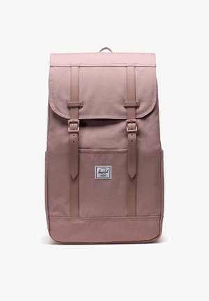 Ružový látkový batoh s horným klopom, dvoma prednými popruhmi a malou značkovou nášivkou. Má obdĺžnikový tvar a hladký povrch.