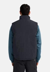 Schwarze gepolsterte Weste mit hohem Kragen, getragen über einem blauen Langarmshirt. Der Rücken hat eine glatte, matte Oberfläche ohne sichtbare Beschläge.
