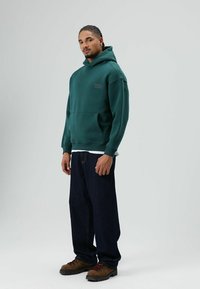 Sweat à capuche vert avec poche avant, marqué "ZERO" sur la poitrine. Associé à un jean large bleu foncé et des bottes de randonnée marron. Design minimaliste.
