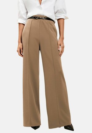 Frau trägt beige hochtaillierte, weit geschnittene Hosen, schwarzen Gürtel mit goldener Schnalle, weißes Hemd mit hochgekrempelten Ärmeln und schwarze spitze Schuhe.