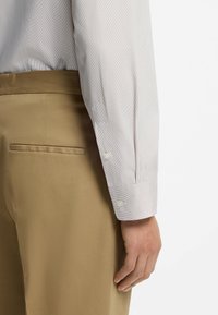 Raidallinen valkoinen paita kalvosin, yhdistetty vaaleanruskeisiin housuihin. Kangas näyttää sileältä; kalvosimessa on nappikiinnitys.