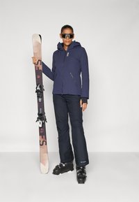 Navy ski jacket with hood, black ski pants, black ski boots, and skis with a pink and tan design. Model wears dark sunglasses.