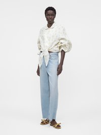 Γυναίκα που φοράει λευκό μπλουζάκι με κίτρινα λουλουδάτα σχέδια δεμένο στη μέση, ανοιχτό μπλε παντελόνι πλατύτερου ποδιού και μπεζ σανδάλια με φιόγκο.