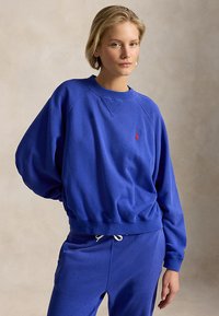 Ξανθιά γυναίκα που φοράει μπλε φούτερ και αντίστοιχο αθλητικό παντελόνι, στέκεται με το ένα χέρι πίσω από την πλάτη της, μπροστά σε μια μονόχρωμη μπεζ επιφάνεια.