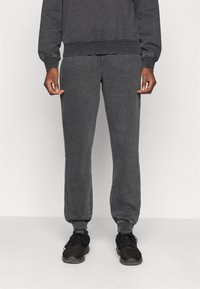 Sive bombažne trenirke imajo ožji kroj, rebraste manšete in stranske šive s kontrastnim šivom. Ujemajo se z črnimi športnimi čevlji.