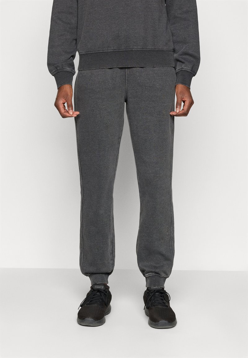 Sive bombažne trenirke imajo ožji kroj, rebraste manšete in stranske šive s kontrastnim šivom. Ujemajo se z črnimi športnimi čevlji.