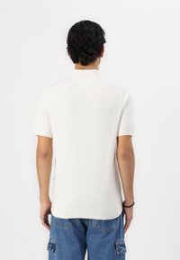 Λευκό κοντομάνικο polo shirt με διακριτή υφή και V-σχήματος λεπτομέρεια στον πίσω λαιμό. Συνδυάζεται με μπλε cargo τζιν.