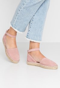 Różowe zamszowe espadryle z paskiem na kostkę, okrągłym noskiem i plecioną platformą z juty. Posiadają płaską podeszwę i przetarte dżinsowe spodnie.