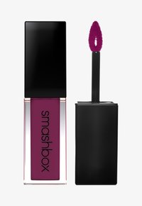 Lápiz labial líquido en un tubo rectangular transparente con una tapa negra brillante. El producto es de color ciruela oscuro y tiene un aplicador de felpa.