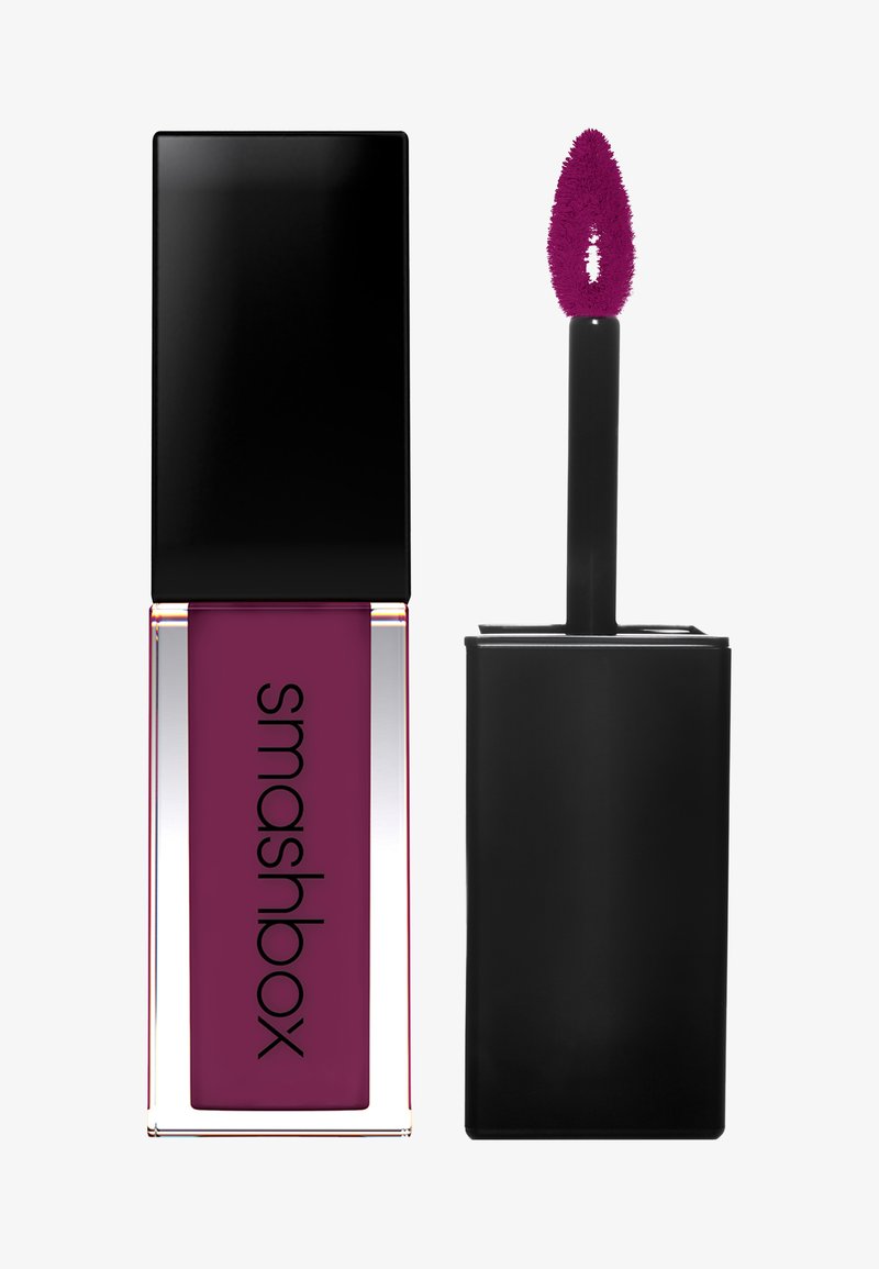 Lápiz labial líquido en un tubo rectangular transparente con una tapa negra brillante. El producto es de color ciruela oscuro y tiene un aplicador de felpa.