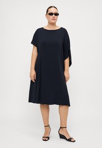 Kjole med korte ermer i A-linjeform laget av ensfarget marineblått stoff, med bred utringning og flytende silhuett. Kombinert med svarte høye sandaler.