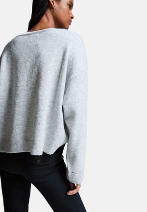 Kvindelig iført lysegrå langærmet sweater og sorte bukser, vist fra ryggen stående mod en enkel hvid baggrund.