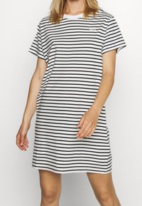 Nainen, jolla on lyhythihainen, polven yläpuolelle ulottuva valkoinen mekko mustilla vaakaraitojen kanssa, seisoo yksinkertaisen valkoisen taustan edessä.