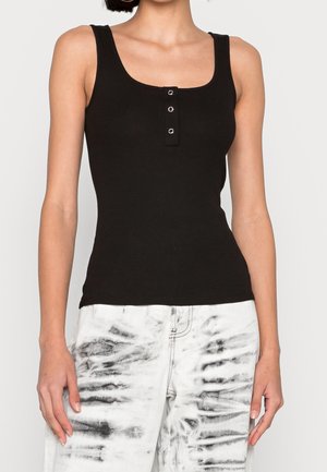 Vrouw draagt een zwart mouwloos geribd topje met drie drukknoopjes aan de voorkant en witte tie-dye broek tegen een effen lichte achtergrond.