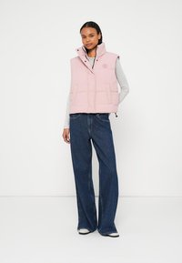 Světlá růžová zateplená vesta s vysokým límcem a cvočky, která má dvě přední kapsy. Nositelná přes šedé tričko s dlouhým rukávem a modré džíny s širokými nohavicemi.