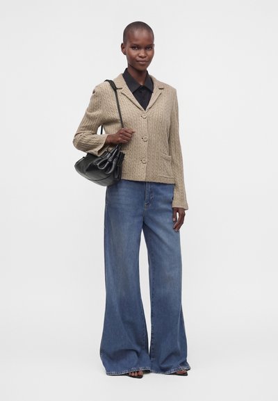Femme portant un blazer en maille beige, une chemise noire, un jean bleu à jambes larges, tenant un sac à épaule noir, debout devant un fond uni.