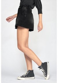 Osoba v černých vysokých teniskách, černé džínové mini sukni a černém topu s dlouhým rukávem, zobrazená od poloviny stehen dolů při chůzi.