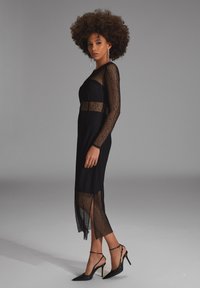 Schwarzes Spitzenkleid mit langen Ärmeln, durchsichtigen Einsätzen und taillierter Silhouette, mit Schlitz am Saum. Kombiniert mit schwarzen High-Heels.