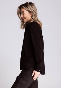 Schwarzes, langärmliges Hemd mit Knöpfen, aus weichem Material, mit spitzem Kragen und abgerundetem Saum, getragen mit schwarzen, gemusterten Hosen.