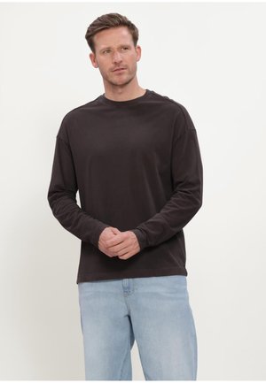 Mann trägt ein dunkles Langarmhemd und hellblaue Jeans, steht mit verschränkten Händen und schaut leicht zur Seite vor einem schlichten Hintergrund.