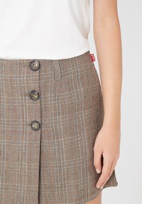 Mujer con una falda de cuadros marrón con tres botones delanteros y una blusa blanca, con la mano descansando a su lado.