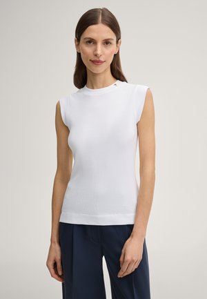 Kobieta o prostych brązowych włosach, ubrana w biały, bezrękawnik i granatowe spodnie, stojąca na tle jasnego, jednolitego tła.