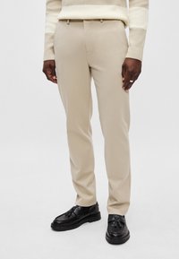 Beige suorat housut, joissa on sileä pinta ja kapea istuvuus. Ominaisuuksiin kuuluvat vyölenkit ja sivutaskut, yhdistettynä mustiin kenkiin.