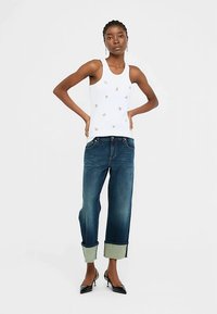 Bela majica brez rokavov z majhnimi zlatimi poudarki, kombinirana z modrimi jeans hlačami z zavihanimi robovi v kontrastni svetlo modri barvi. Črne čevlje z visokimi petami.