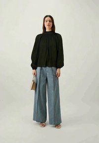 Blusa preta de mangas compridas com decote alto, combinada com calças de ganga azul claro de perna larga e sandálias beges, segurando uma pequena mala castanha.