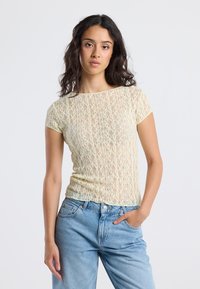 Woman with long dark hair wearing a sheer yellow lace short-sleeve top and light blue high-waisted jeans, posing with one hand in pocket.