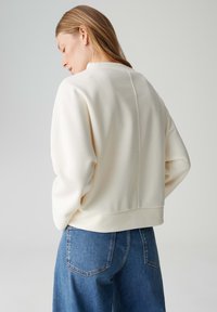 Cr�èmekleurige, zachte sweatshirt met een ontspannen pasvorm, verlaagde schouders en een cropped zoom. Gecombineerd met hooggesneden blauwe spijkerbroek.