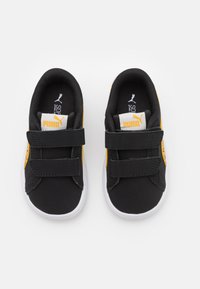 Svarta och gula sneakers med en slät textur, som har två kardborreband, vita sulor och en logotyp på tungan. Dessutom finns det en sidologotyp.