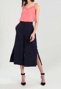 Camisole en satin rose avec bordure en dentelle associée à un pantalon culotte taille haute bleu marine avec fentes latérales. Des talons à bride à la cheville noirs complètent le look.