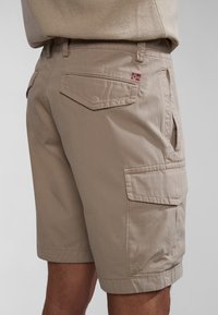 Pantalones cortos cargo beige hechos de tela ligera, con múltiples bolsillos, detalles de costura y un pequeño acento de bandera en la parte trasera.
