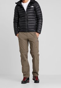 Czarna puchowa kurtka z zamkiem błyskawicznym, zestawiona z beżowymi spodniami typu cargo z kieszeniami na zamek oraz czarnymi butami trekkingowymi z czerwonymi akcentami.