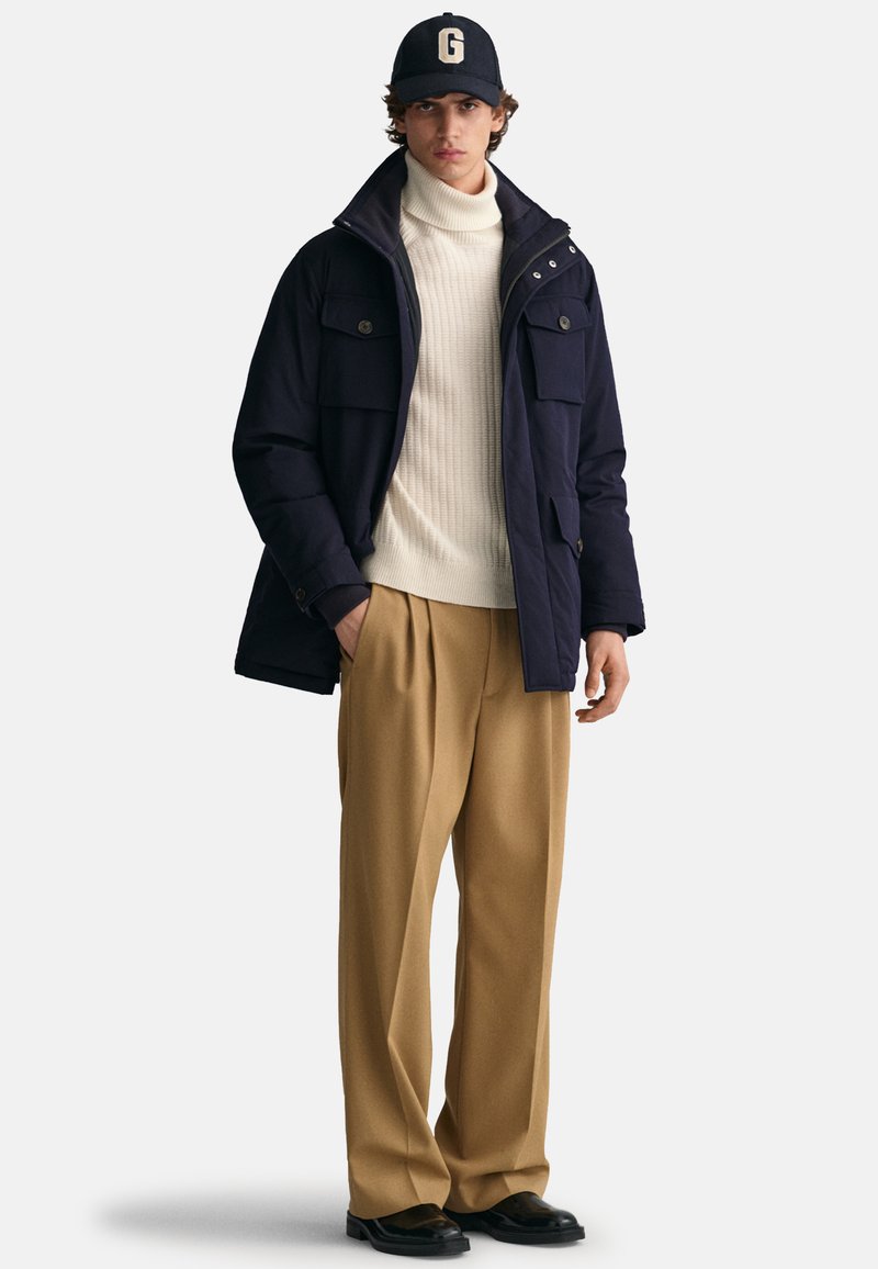 Giacca isolata blu marina con grandi tasche, dolcevita crema, pantaloni pieghettati color beige e un berretto nero con un logo bianco "G". Scarpe nere.