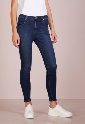 Persoon gekleed in donkerblauwe strakke jeans, witte sneakers en een wit mouwloos topje, staand tegen een neutrale achtergrond.